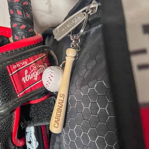 May include: A baseball-themed keychain featuring a miniature wooden bat with the word CARDINALS engraved on it, attached to a small white baseball charm. The keychain is connected to a silver metal ring and a rectangular silver tag with text.