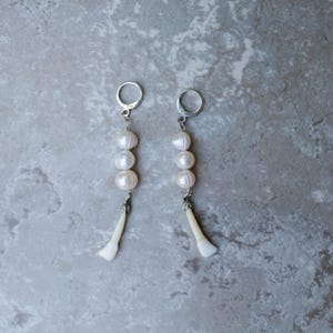 May include: A pair of dangle earrings featuring three white pearls and a white, tooth-shaped charm. The earrings have silver-colored ear wires and are displayed against a gray, mottled background.