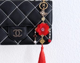 May include: A black quilted handbag with a decorative red flower charm. The charm features a red flower, gold accents, a butterfly, and a red tassel. The handbag has a chain strap and a silver logo.