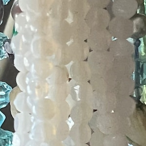 May include: Close-up of a beaded ornament with a variety of beads. The ornament features strands of white, light blue, and iridescent beads. Some beads have a metallic finish, and others are clear.