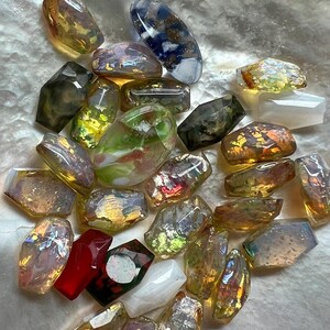 May include: A collection of iridescent, faceted glass beads in various shapes and sizes. The beads display a range of colors, including gold, green, red, and blue, with a shimmering, reflective quality. Some beads are rectangular, while others are more organic in shape.