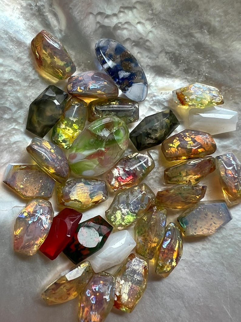 May include: A collection of iridescent, faceted glass beads in various shapes and sizes. The beads display a range of colors, including gold, green, red, and blue, with a shimmering, reflective quality. Some beads are rectangular, while others are more organic in shape.