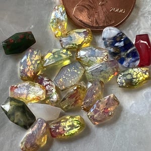 May include: A collection of iridescent glass beads in various shapes and colors, including yellow, red, and green. A copper penny with the year 2000 is in the background, providing scale. These beads are suitable for jewelry making and crafts.