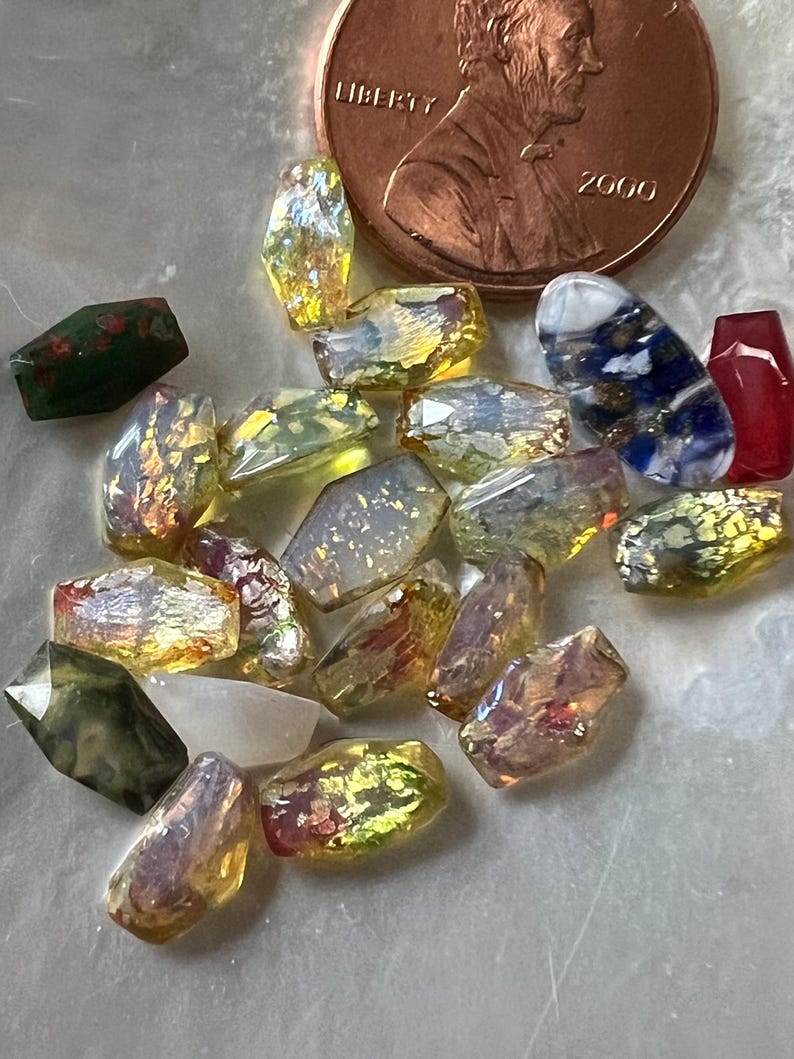 May include: A collection of iridescent glass beads in various shapes and colors, including yellow, red, and green. A copper penny with the year 2000 is in the background, providing scale. These beads are suitable for jewelry making and crafts.