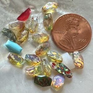 May include: A collection of small, faceted gemstone beads in various colors, including yellow, red, and iridescent hues. A copper penny is included for size reference. The beads are irregularly shaped and appear to be tumbled.