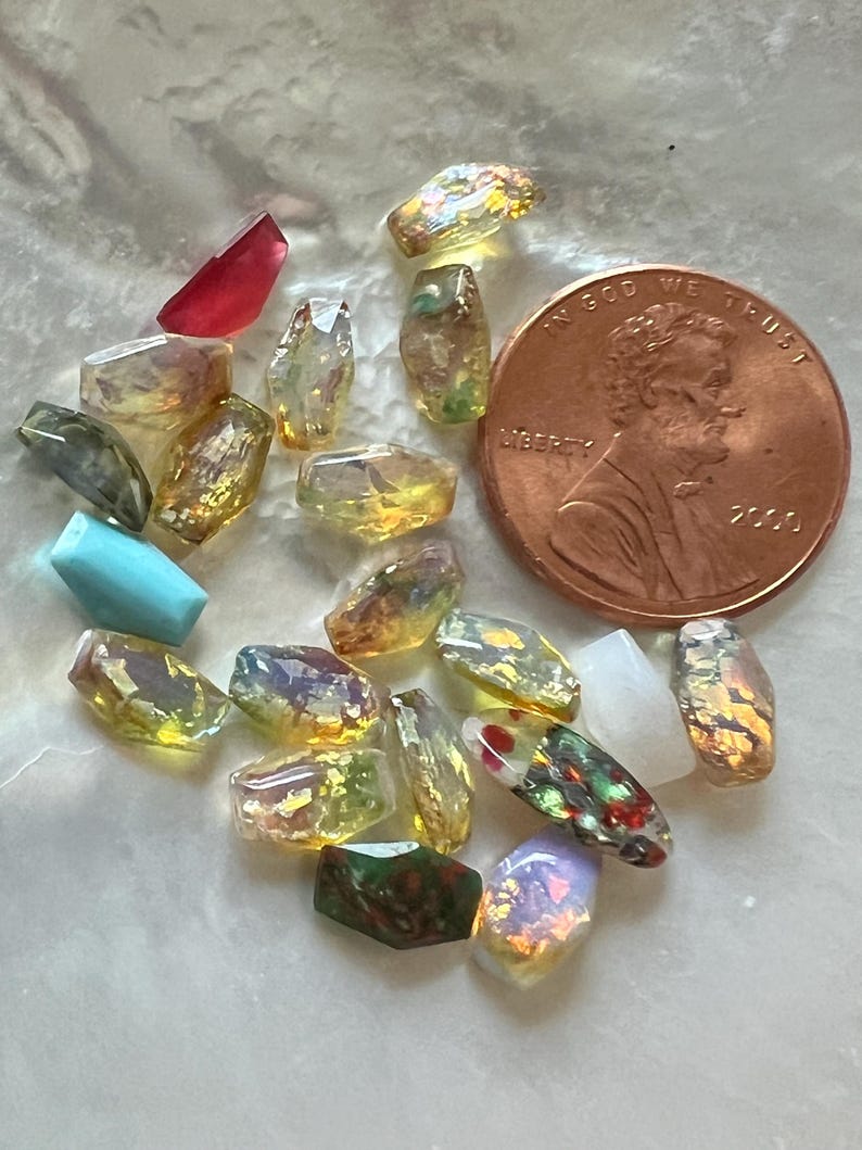 May include: A collection of small, faceted gemstone beads in various colors, including yellow, red, and iridescent hues. A copper penny is included for size reference. The beads are irregularly shaped and appear to be tumbled.