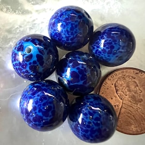 May include: Six round, dark blue glass beads with a speckled pattern in a lighter shade of blue. Each bead has a hole for stringing. A copper penny is next to the beads for size comparison.
