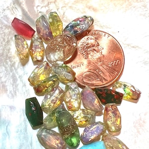 May include: A collection of colorful, iridescent beads in various shapes and sizes, including faceted and oval designs. The beads are in shades of yellow, green, purple, and red. A copper penny is also present, with the year 2000 visible.
