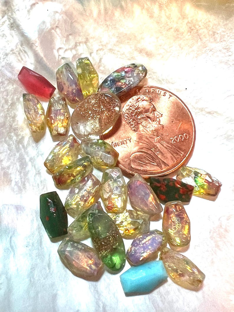 May include: A collection of colorful, iridescent beads in various shapes and sizes, including faceted and oval designs. The beads are in shades of yellow, green, purple, and red. A copper penny is also present, with the year 2000 visible.