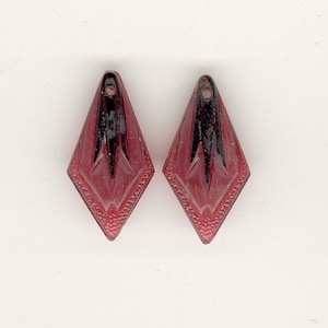 May include: Two red and black diamond-shaped earrings. Each earring has a black central design and a decorative border. The earrings have a small hole at the top for hanging. The earrings are likely vintage or antique.