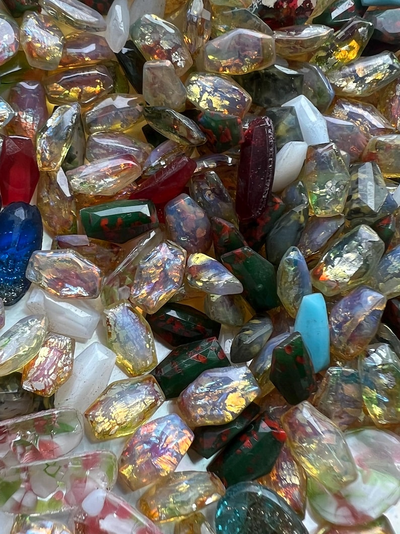 May include: A collection of polished gemstones in various colors, including red, blue, green, and clear. The stones have an iridescent quality, with flashes of gold and other colors. The gemstones are irregularly shaped and appear to be tumbled.