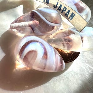 May include: Close-up of several translucent, teardrop-shaped glass beads with white and purple swirls. A tag that reads "MADE IN JAPAN" is visible. The beads are resting on a soft, white surface.
