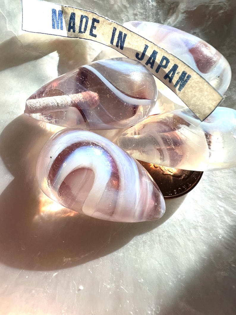May include: Close-up of several translucent, teardrop-shaped glass beads with white and purple swirls. A tag that reads "MADE IN JAPAN" is visible. The beads are resting on a soft, white surface.