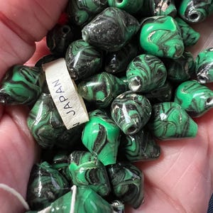 May include: A collection of green and black swirled beads. The beads are a mix of shapes and sizes, with a glossy finish. A tag that says "JAPAN" is visible. The beads are strung together with white string.