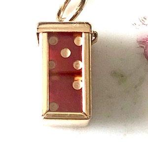 May include: A gold-colored pendant in the shape of a domino. The domino has a red background with white dots. The pendant has a loop at the top for attaching to a chain or bracelet. The domino shows the number five.