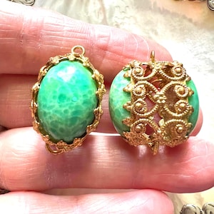 May include: Two vintage-style pendants. One has a gold-colored filigree frame around a green, oval-shaped stone. The other pendant features a gold filigree design around a round, green stone. Both have a loop for hanging.