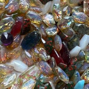 May include: A collection of colorful, iridescent glass beads and stones. The assortment includes various shapes and sizes, with colors ranging from clear and gold to red, green, and blue. Some stones have a speckled pattern.