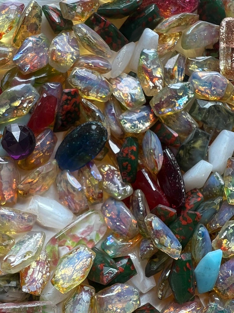 May include: A collection of colorful, iridescent glass beads and stones. The assortment includes various shapes and sizes, with colors ranging from clear and gold to red, green, and blue. Some stones have a speckled pattern.
