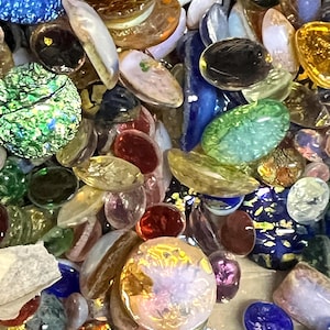 May include: A collection of assorted glass cabochons in various shapes and sizes, featuring a mix of colors including deep blue, emerald green, amber, clear, and iridescent finishes with some pieces displaying gold foil flecks and textures.