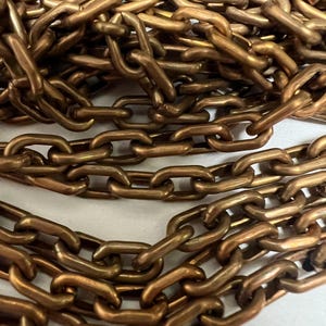 May include: Close-up of a pile of bronze-colored metal chains. The chains are linked together, creating a textured surface. The links are oval-shaped and have a slightly worn appearance, suggesting age or use. The chains are arranged in a haphazard manner.