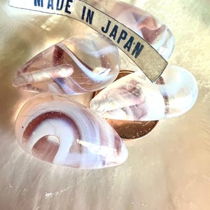 May include: Four teardrop-shaped glass beads with white, pink, and purple swirls. A tag that reads "MADE IN JAPAN" is draped across the beads. The beads are on a light pink surface.