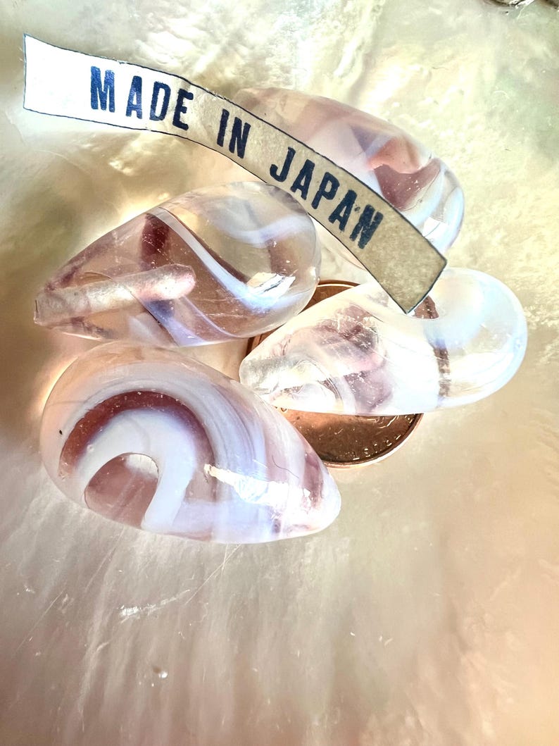 May include: Four teardrop-shaped glass beads with white, pink, and purple swirls. A tag that reads "MADE IN JAPAN" is draped across the beads. The beads are on a light pink surface.