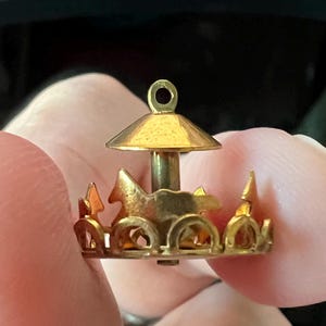 May include: A gold-toned metal charm shaped like a miniature carousel. The charm features a central pillar, a conical roof with a small loop at the top for attachment, and a circular base decorated with cutout silhouettes of horses and arches.