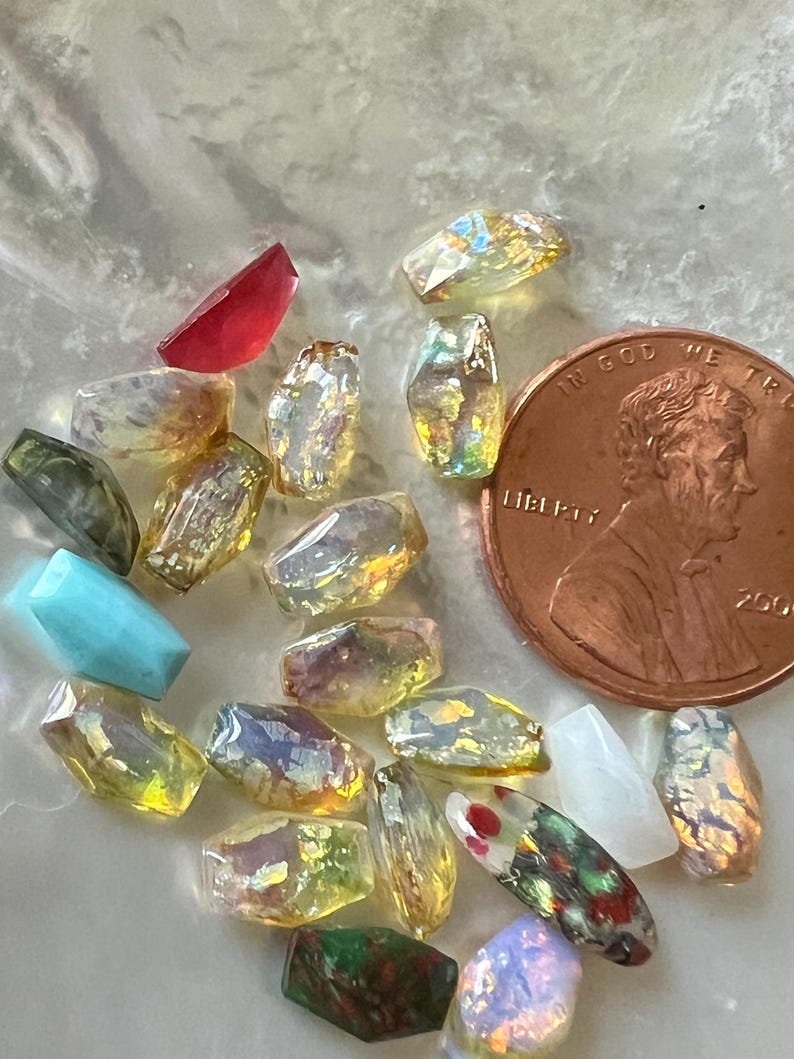 May include: A collection of various gemstone beads in shades of yellow, green, red, and blue. The beads are faceted and have an iridescent quality. A US penny is included for size reference.