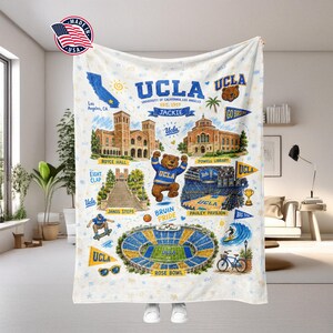 May include: A white fleece blanket featuring a UCLA theme with illustrations of Royce Hall, Powell Library, Janss Steps, Pauley Pavilion, and the Rose Bowl. It includes a bear mascot, the text UCLA, Bruin Pride, and Jackie, plus sports icons.
