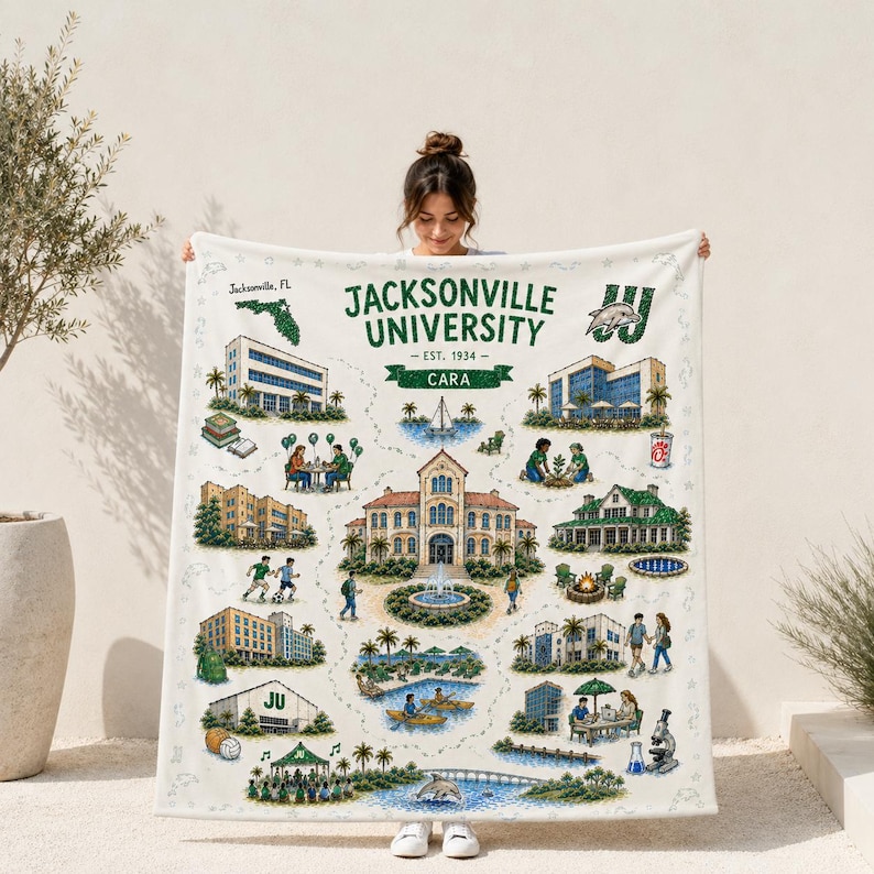 May include: A white fleece blanket featuring a detailed illustration of the Jacksonville University campus. The design includes various buildings, a fountain, palm trees, students, a dolphin mascot, and the text Jacksonville University Est. 1934.