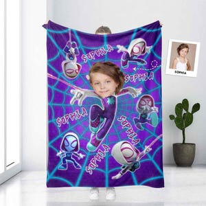 Puede incluir: Una manta morada con temática de Spider-Man con una cara personalizada y el nombre "SOPHIA" repetido. La manta tiene un diseño de telaraña y múltiples ilustraciones de personajes de Spider-Man en varias poses. Una pequeña foto de una niña está en la esquina superior derecha.