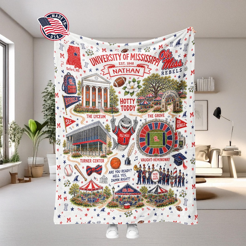 May include: A white fleece blanket featuring University of Mississippi graphics in red and blue. Illustrations include the Lyceum, Vaught-Hemingway Stadium, a shark mascot, a marching band, and text such as Hotty Toddy, Nathan, and Ole Miss Rebels.