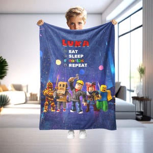 May include: A blue blanket featuring the name "LUKA" in red letters, with the words "EAT, SLEEP, ROBLOX, REPEAT" and cartoon characters. The blanket has a night sky background with colorful dots and city silhouettes.
