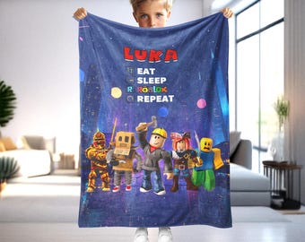 May include: A blue blanket featuring the name "LUKA" in red letters, with the words "EAT, SLEEP, ROBLOX, REPEAT" and cartoon characters. The blanket has a night sky background with colorful dots and city silhouettes.