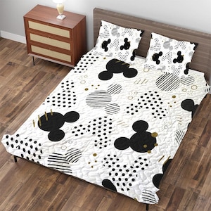 Puede incluir: Juego de cama blanco con un estampado de Mickey Mouse en negro y dorado. El juego incluye un edredón y dos fundas de almohada. El edredón presenta un patrón repetido de siluetas de Mickey Mouse, lunares y rayas.