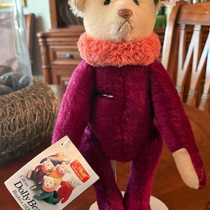 May include: A Steiff Circus Dolly Bear replica 1913 with a cream-colored head, a bright coral-colored ruffled neck collar, and a deep magenta-colored body. The bear features a small metal tag in its ear and a white product tag attached.