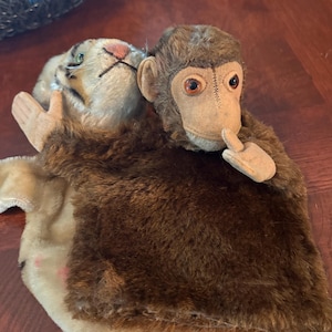 May include: A plush hand puppet featuring a brown monkey with a tan face and a finger held to its mouth, layered over a white and black striped tiger puppet. The monkey has dark brown fur and orange eyes, while the tiger has green eyes and a pink nose.