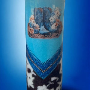 May include: A tall, cylindrical tumbler with a blue cowboy boot design. The tumbler features a cowhide pattern at the bottom, a blue band with glitter, and a clear top. The boots are light blue with floral accents.