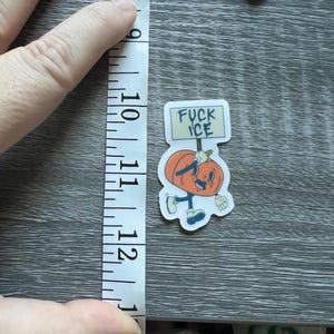 May include: A sticker featuring a cartoon pumpkin character holding a sign that reads "FUCK ICE." The pumpkin is orange with a smiling face, wearing blue shoes and gloves. The sticker measures approximately 2.5 inches in height.