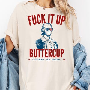 May include: A cream-coloured t-shirt featuring the text "FUCK IT UP" in red above an illustration of George Washington wearing sunglasses in the style of the American flag. The word "BUTTERCUP" is below the illustration in red. The text "1776 ENERGY. 2025 PROBLEMS." is below.