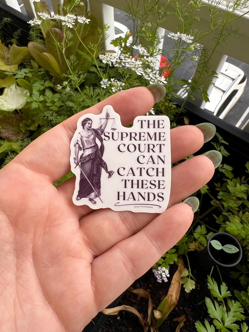 Op de afbeelding: Een witte sticker met een zwart-witte illustratie van een vrouw die een weegschaal vasthoudt. De tekst op de sticker luidt "The Supreme Court Can Catch These Hands."