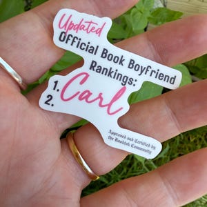 May include: A white die-cut sticker with black and pink text that reads: Updated Official Book Boyfriend Rankings: 1. Carl 2. Approved and Certified by the Booktok Community. The sticker features a mix of script and sans-serif fonts.