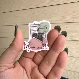 May include: A sticker with a pink and green background featuring a black and white illustration of a guillotine with the text "Marie said hi" in pink.