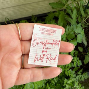 Può includere: Una scheda della biblioteca bianca e rossa con il testo "Overstimulated but Well Read" in corsivo rosso. La scheda include anche il testo "Si prega di restituire questo libro nelle stesse condizioni in cui te ne sei innamorato."