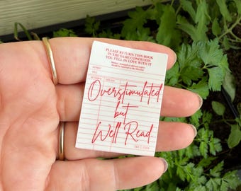 May include: A white and red library card with the text "Overstimulated but Well Read" in red script. The card also includes the text "Please return this book in the same condition you fell in love with it."