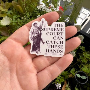 Op de afbeelding: Een witte sticker met een zwart-witte illustratie van de figuur van Justitia die een weegschaal en een zwaard vasthoudt. De tekst op de sticker luidt "The Supreme Court Can Catch These Hands."