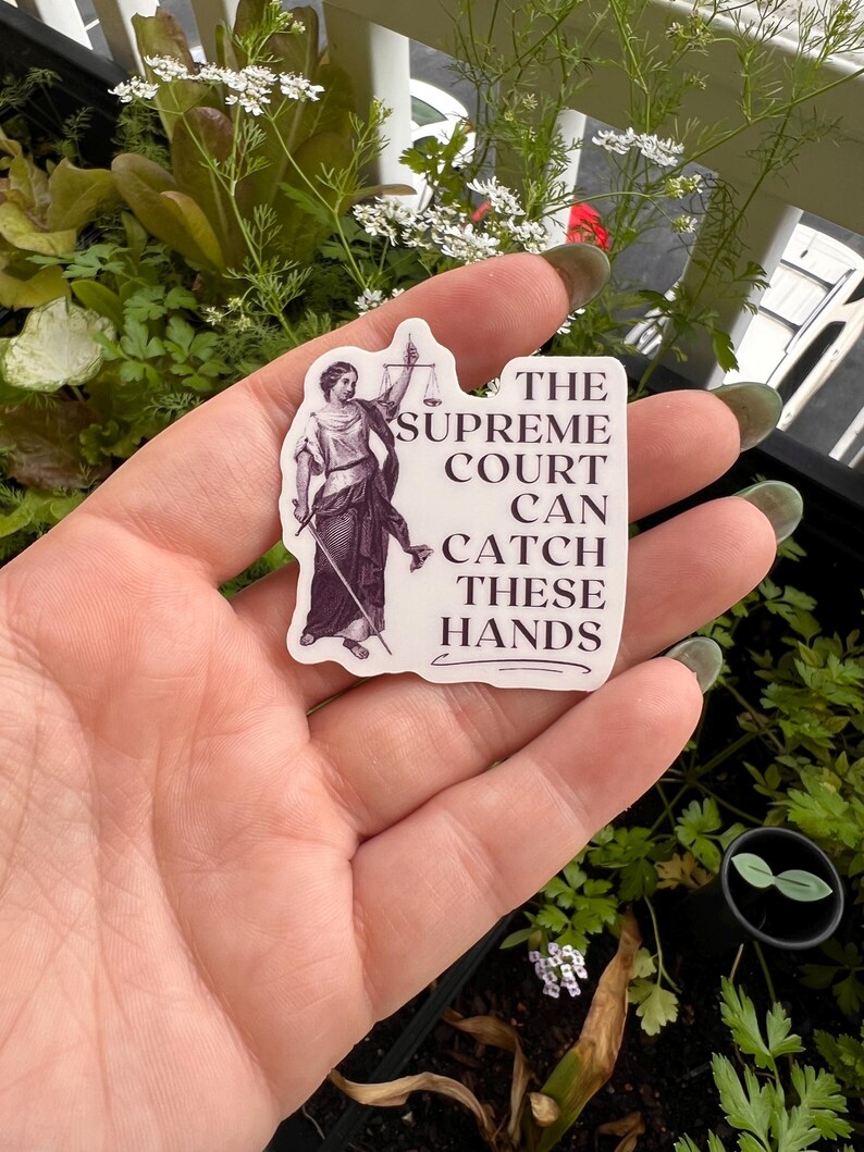 Op de afbeelding: Een witte sticker met een zwart-witte illustratie van de figuur van Justitia die een weegschaal en een zwaard vasthoudt. De tekst op de sticker luidt "The Supreme Court Can Catch These Hands."