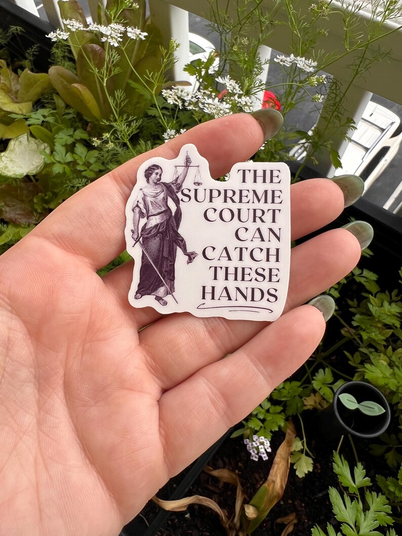 Op de afbeelding: Een witte sticker met een zwart-witte illustratie van de figuur van Justitia die een weegschaal en een zwaard vasthoudt. De tekst op de sticker luidt: "The Supreme Court Can Catch These Hands."