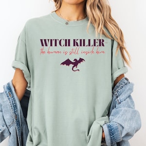 Può includere: T-shirt verde chiaro con la scritta "WITCH KILLER - the human is still inside him" in bordeaux. Una grafica di drago viola è sotto il testo. La maglietta è indossata con una giacca di jeans.