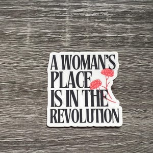 May include: A white sticker with the text "A Woman's Place Is In The Revolution" in a bold, black font. Two red flower illustrations are on the right side of the sticker. The sticker is on a grey wood-grain surface.